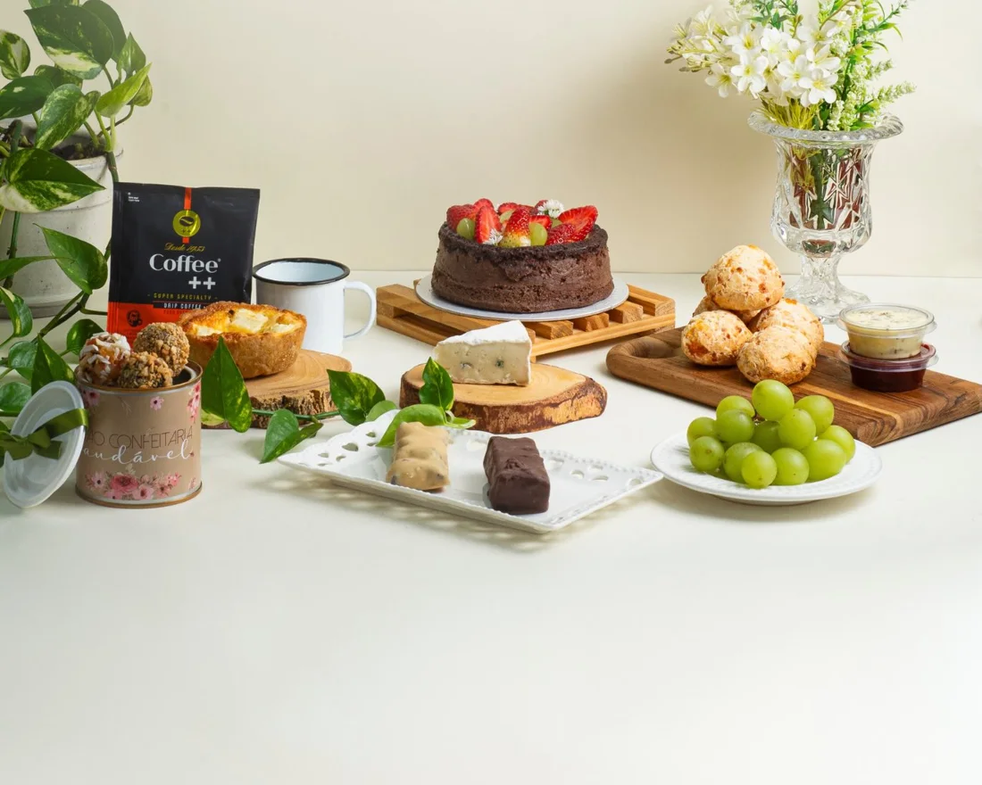 Mesa posta com bolo de chocolate com morangos, pães de queijo, frutas e flores da Grão